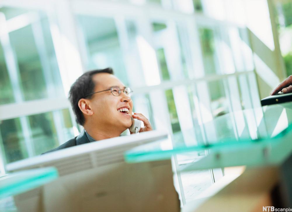 En mann smiler mens han snakker i telefonen foran en datamaskin. Foto. 