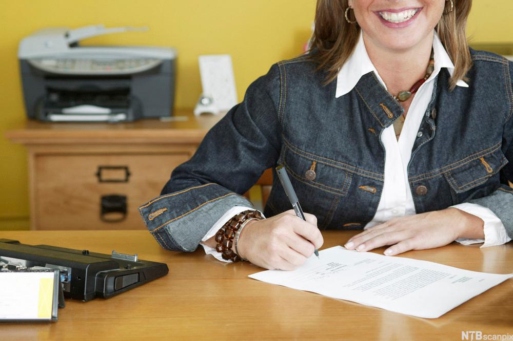 Utsnitt av ei smilande kvinne som sit ved ein pult på eit kontor og signerer ein kontrakt. Foto.