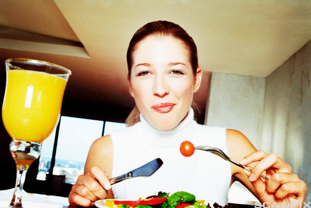 En kvinne sitter ved et bord og spiser salat og drikker appelsinjus. Foto.