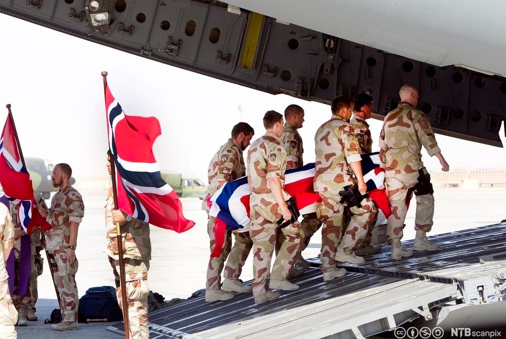 Seks soldater bærer ei kiste med norsk flagg på inn i et fly. Ved siden av står soldater med norske flagg til ære for den falne. Foto.