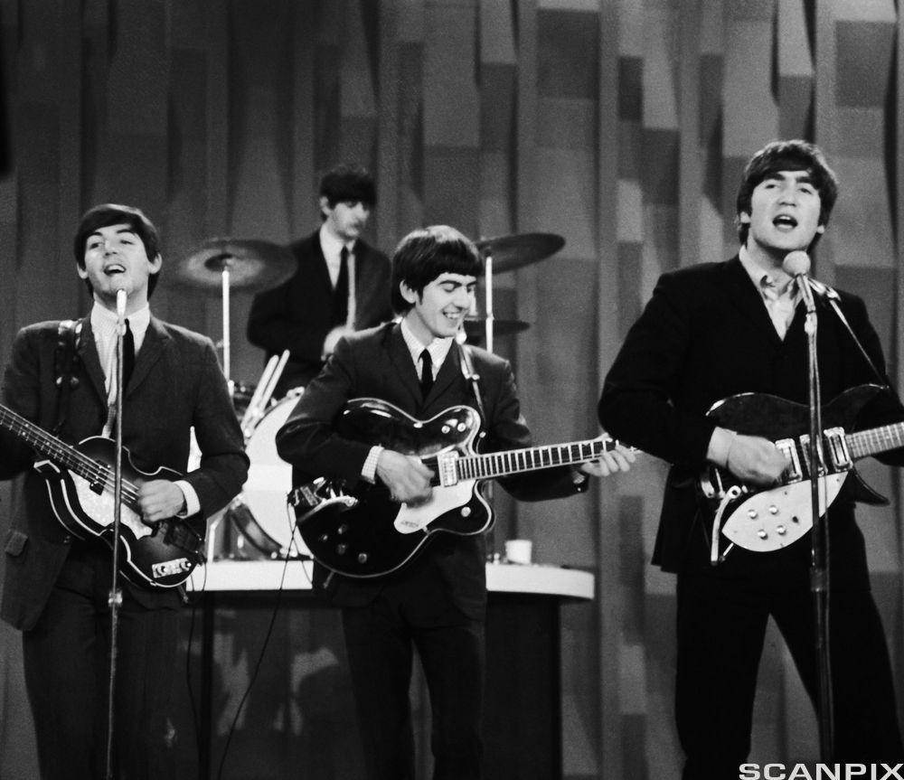 Photo: The Beatles in concert in the early 1960s. All four members of the band are in the picture. They are wearing suits and ties.