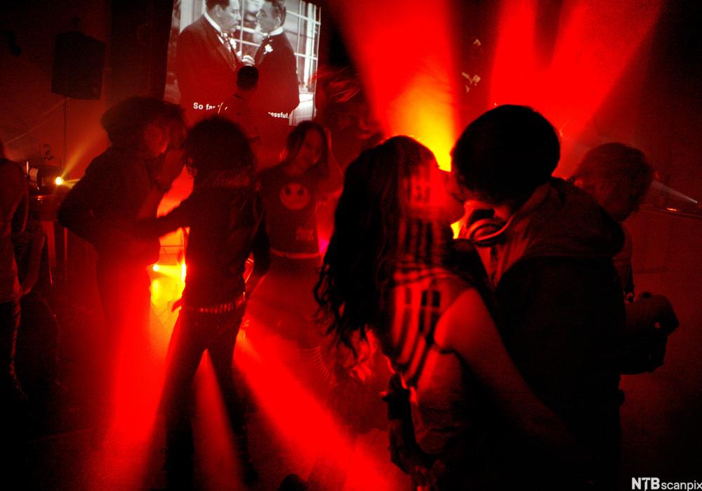 Ungt par som kysser på fest. De står på et dansegulv, og rødt lys flommer utover rommet. Mange andre par danser i bakgrunnen, og det er et lerret med en sort-hvitt-film på veggen. Foto.