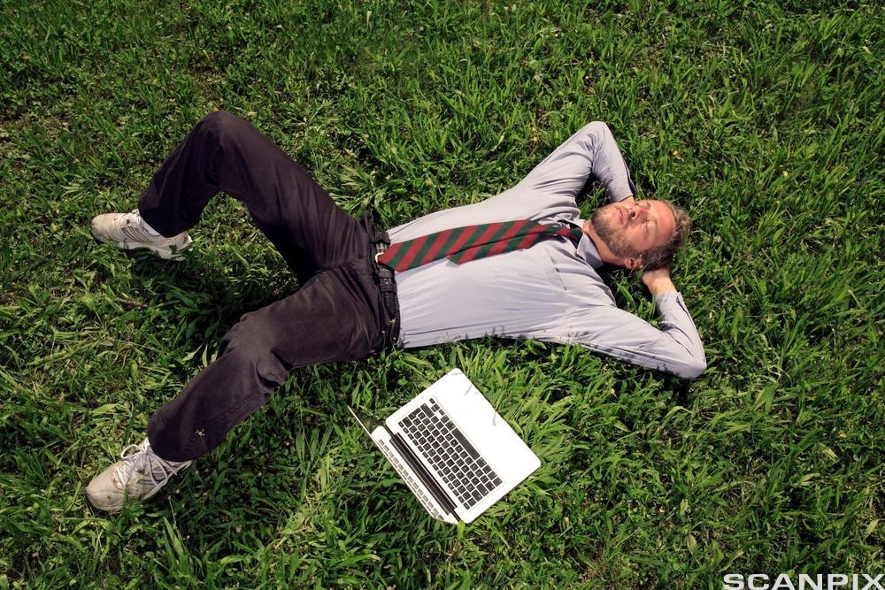 Mann med skjorte og slips ligger i gresset og lukker øynene. Det er en bærbar pc ved siden av ham. Foto.