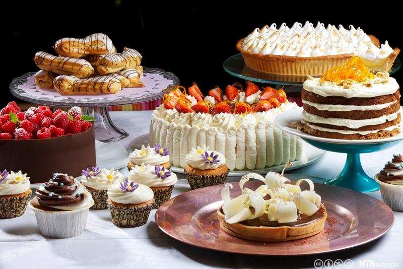 Mange forskjellige forseggjorte kaker på et kakebord, blant annet jordbærbløtkake, gulrotkake, muffins med smørkrem, vannbakkels og en sitronterte. Foto.