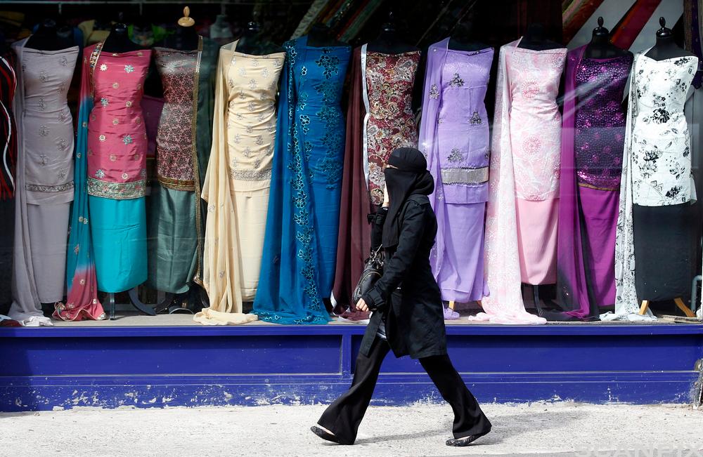 Woman wearing a niqab and dressed in black passes by a window display of colourful Indian saris. Photo. 