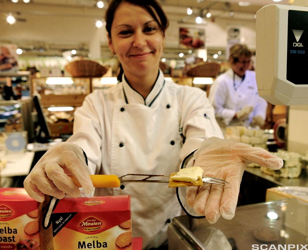 En smilende kvinne i kokkejakke deler ut smaksprøver på ost og kjeks i en matbutikk. Foto.