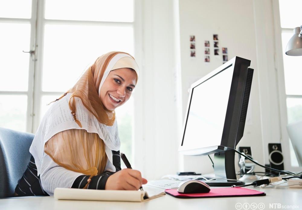 Kvinne med hijab sitter ved et arbeidsbord med datamaskin og noterer. Foto.