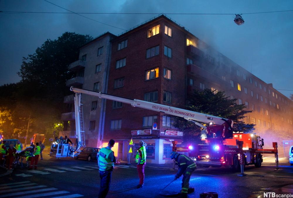 Situasjonsbilete av brann i ei boligblokk. Politi og brannmannskapar i refleksvestar, og en lift. Foto.