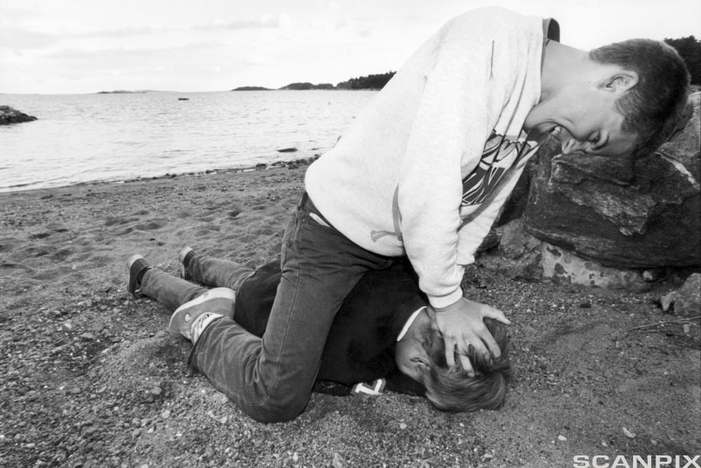 Ein gut sit oppå ein annan gut, og pressar ansiktet hans ned i sanda på ei strand. Foto.