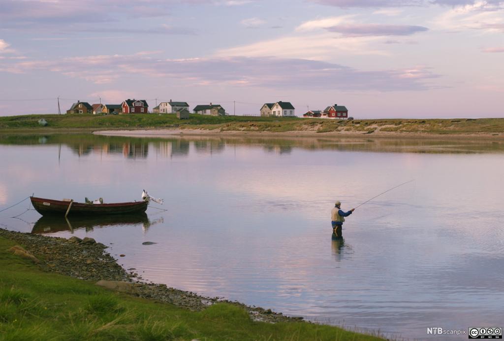 En mann som driver fluefiske på stille vann. En båt er fortøyd ved siden av ham, og det er hus i bakgrunnen. Foto.