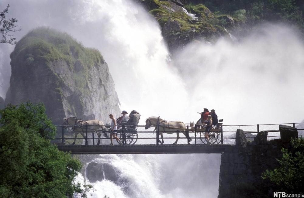 Mennesker kjører hest og kjerre over en smal bro foran en stor, skummende foss. Foto.