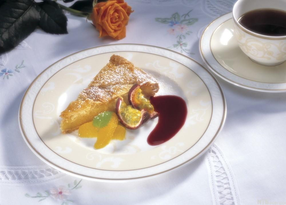 Et stykke fyrstekake på en tallerken, pyntet med appelsinfileter, pasjonsfrukt og rød saus. En kopp med kaffe står ved siden av. Foto.