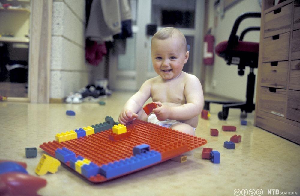 Lite barn i bare bleie sitter på gulvet og leker med duplo og smiler. Foto.
