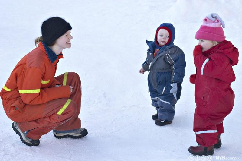 En barnehageansatt sitter på huk foran to små barn og smiler til dem. Alle tre har vinterklær, de er ute i snøen. Foto.