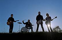Foto av fire musikarar ute i naturen, tre med gitar og ein med trommer.