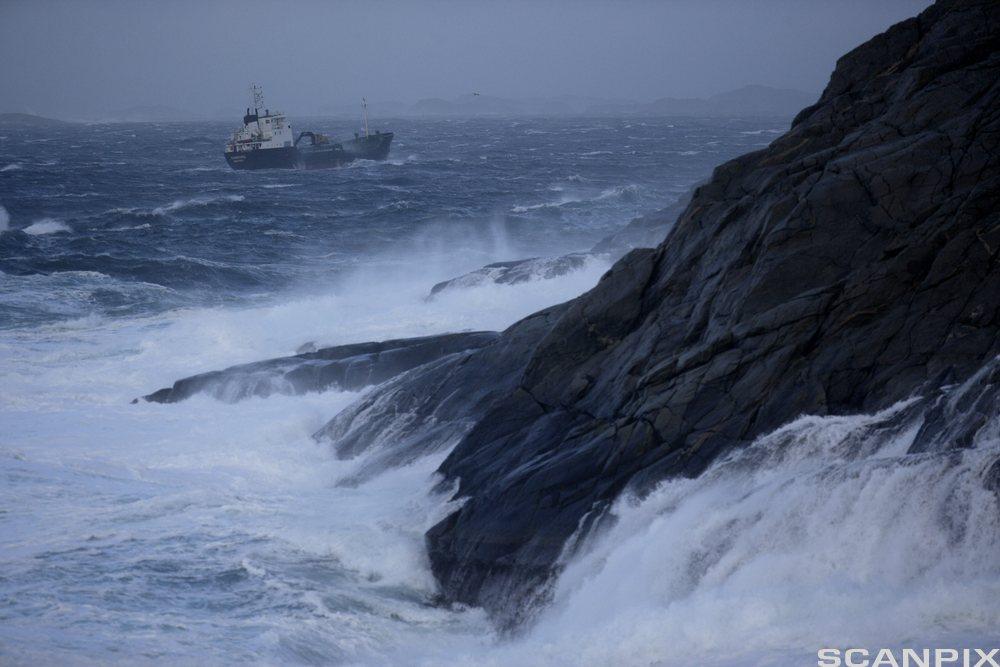 En lastebåt er ute på stormfullt hav. Høye bølger slår mot svaberg framme i bildet. Foto.