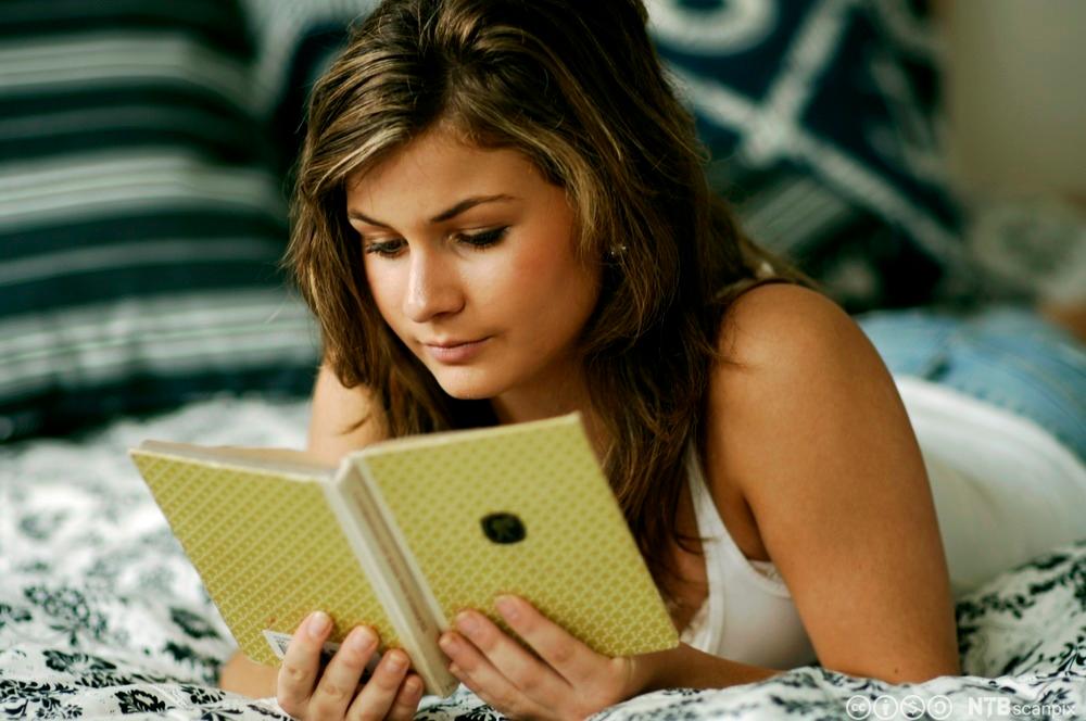 Ung jente ligger på magen i senga og leser i ei bok. Boka har gult omslag, og jenta holder rundt den med begge hendene. Foto.