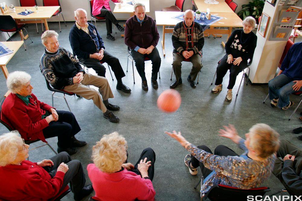 Eldre mennesker sitter på stoler i ring og kaster en ball til hverandre innendørs. Foto.