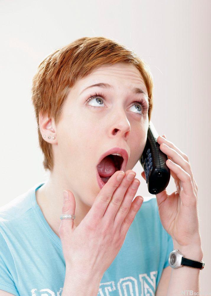 Ung kvinne holder en telefon mot øret og gisper sjokkert. Foto. 