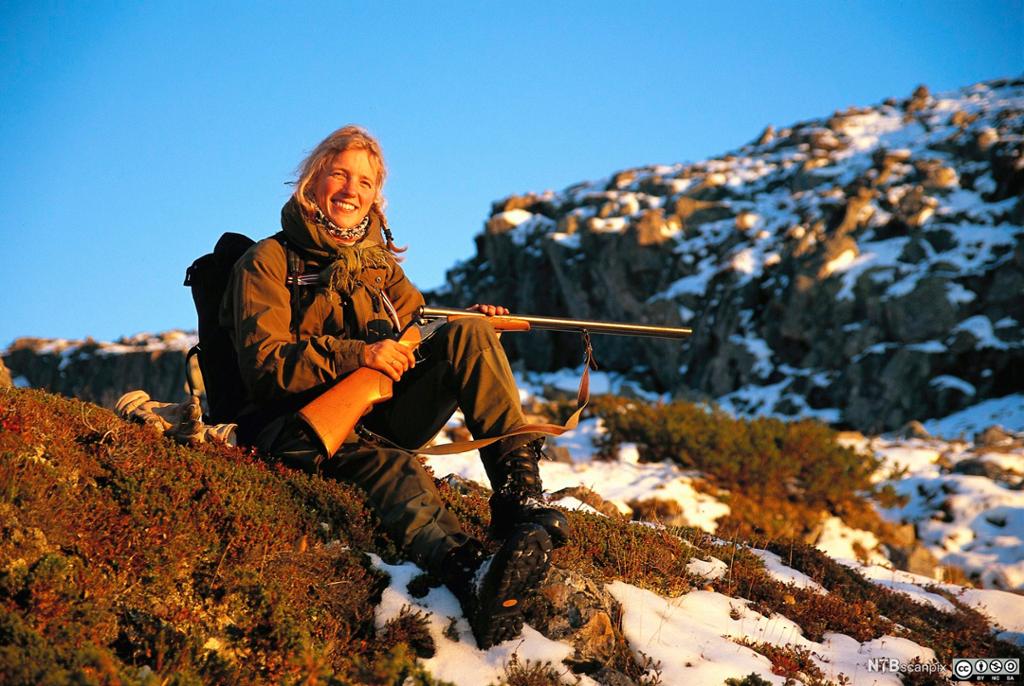 Kvinne sitter i landskap med høstfarga lyng og snøflekker, med jaktvåpen i fanget. Foto.