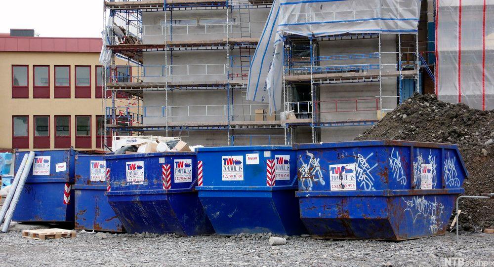 Ei rekke blå konteinarar står framfor eit murbygg under oppføring. Foto.