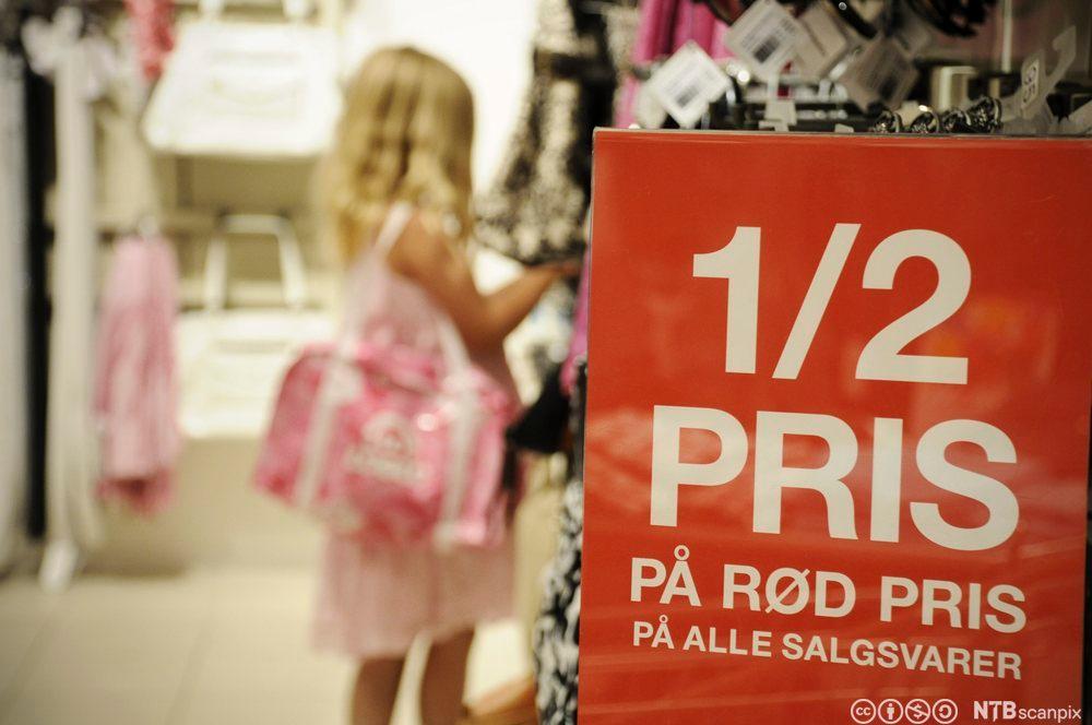 Ei lita jente ser på varer i en butikk. I forgrunnen henger det en rød plakat om salgsvarer til halv pris. Foto.