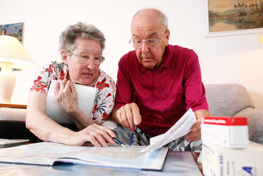 Eldre par sitter i sofaen mens de lener seg fram over et atlas på bordet og peker i det med hver sin penn. Foto.
