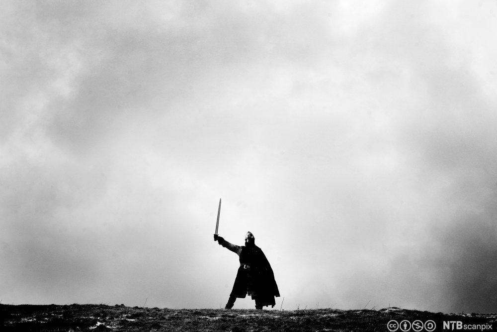 En viking i full utrustning som peker med sverdet opp mot en grå og skyfull himmel. Foto.
