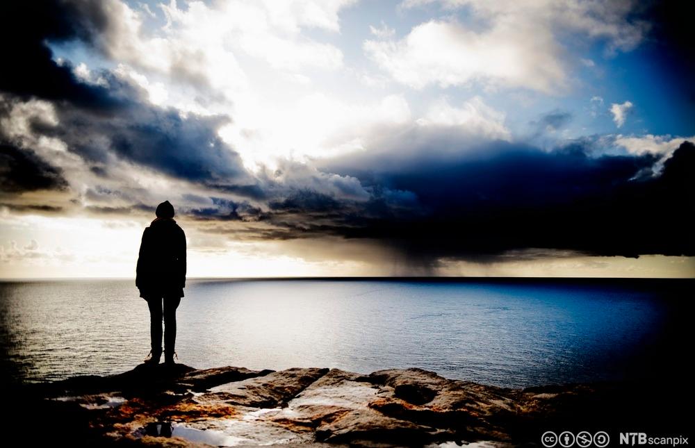 Ein person står på eit svaberg og ser utover havet. Skyer sprekk opp på himmelen. Foto.