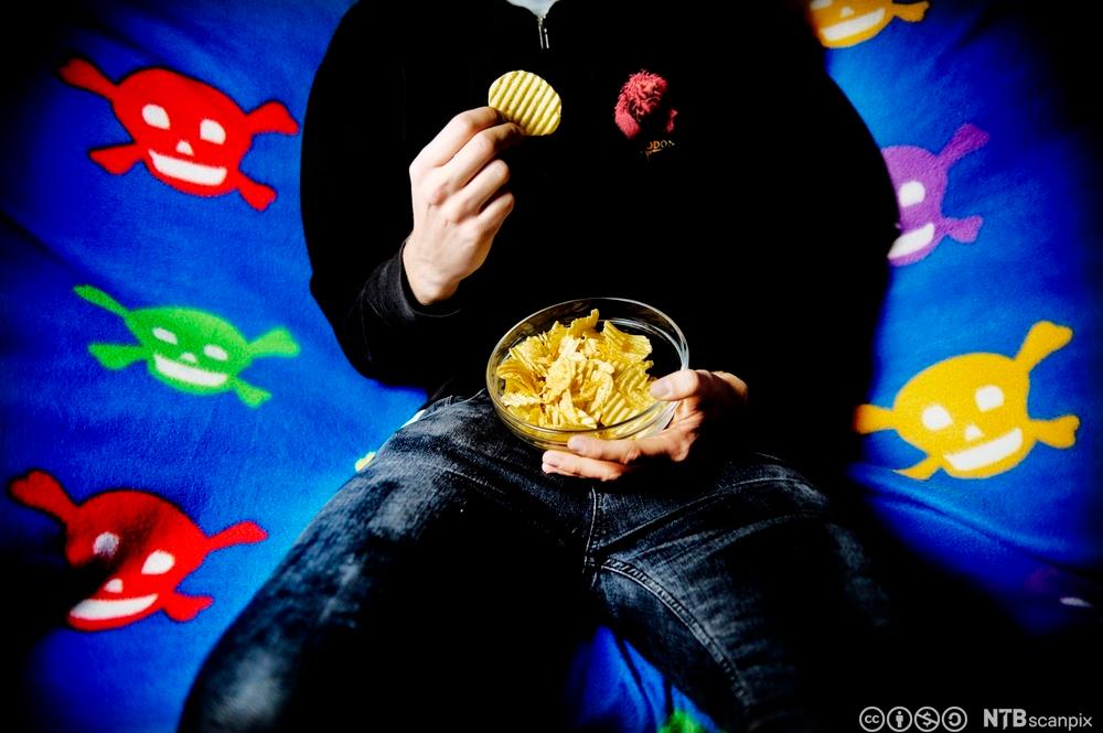 Gutt sitter i sofa og spiser potetgull. Foto