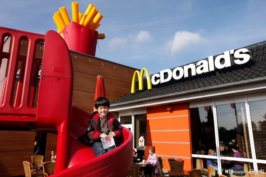 Utsida av en McDonald's-restaurant. Et barn sitter på lekestativet. På taket ser vi McDonald's-logoen. Foto.