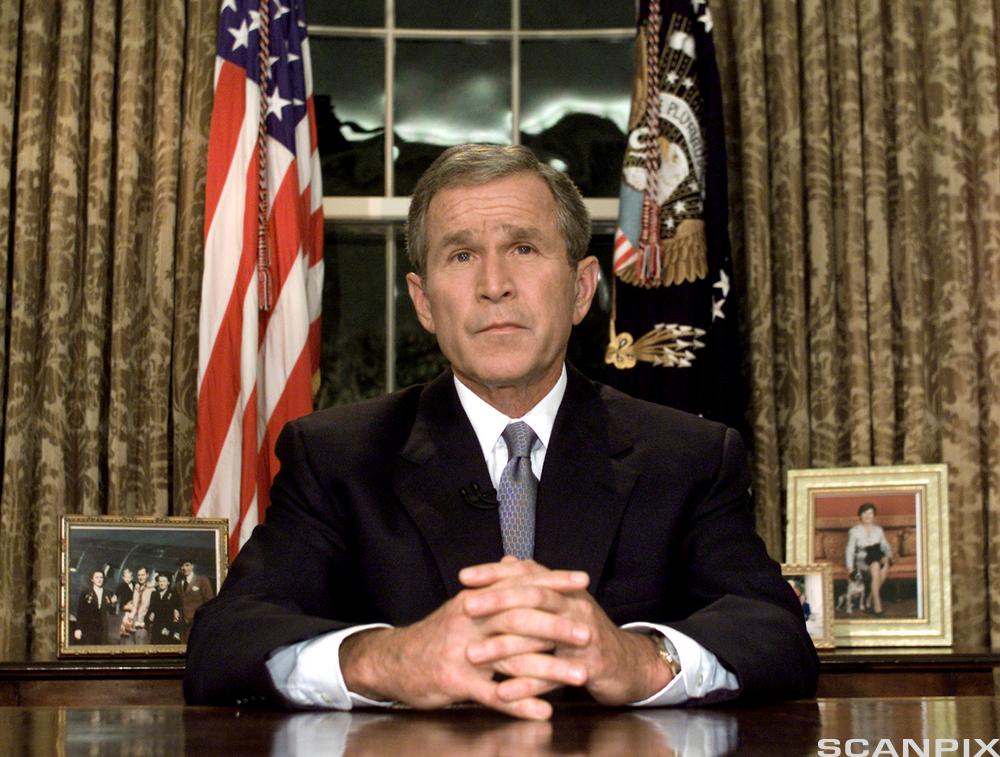 George W. Bush sit ved eit skrivebord med hendene folda føre seg. I bakgrunnen er det eit amerikansk flagg. Det står også meir private familiebilete på benken bak han. Foto.