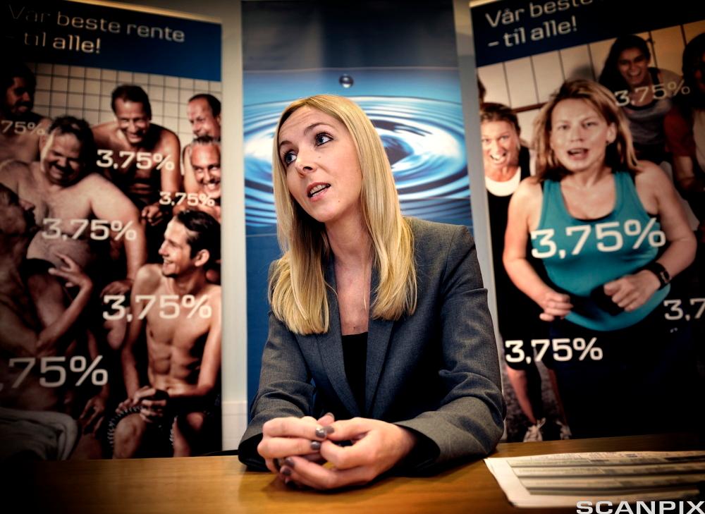 Kvinne ved pult og med plakater med prosenttall på i bakgrunnen. Foto.