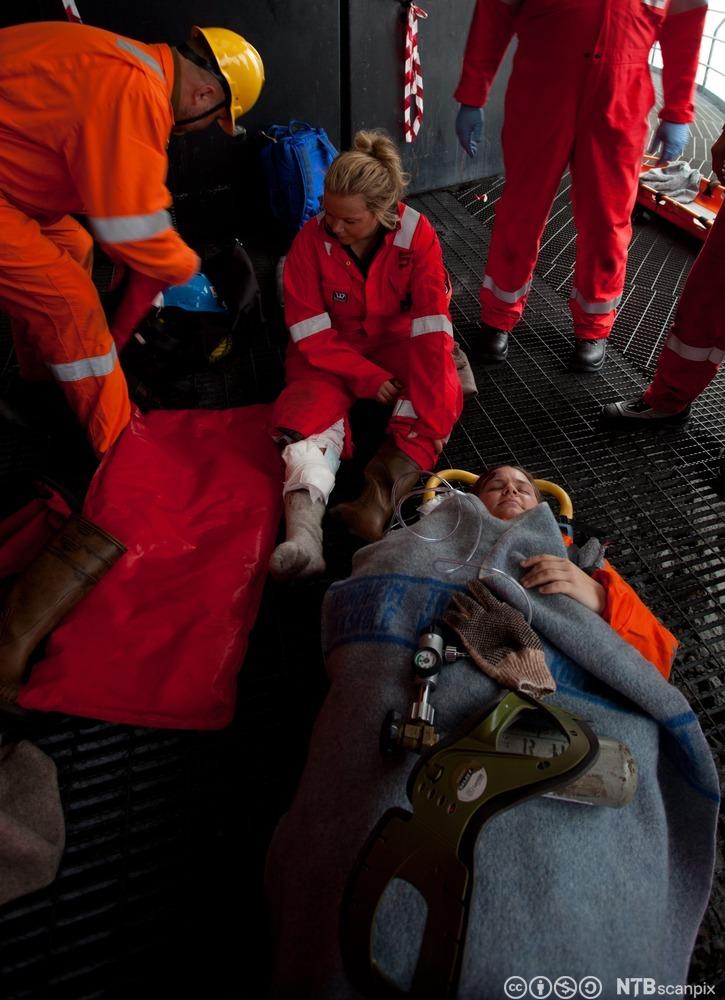 Offshore-arbeidere på førstehjelpskurs. En person ligger på ei båre med et pledd over seg. En kvinne sitter på bakken med bandasje på leggen. Tre personer står rundt dem og hjelper dem. Foto.