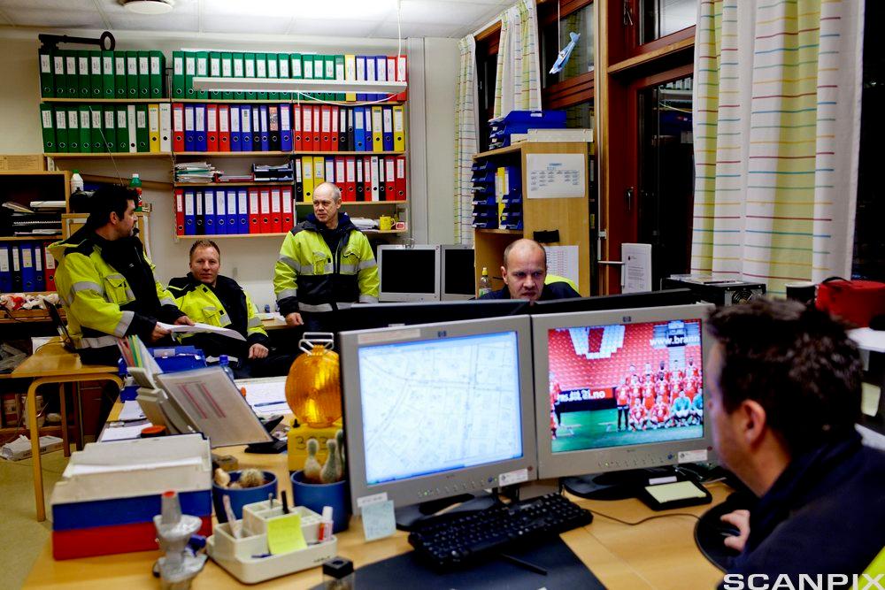En gruppe menn i arbeidstøy sitter i samtale på et kontor. I forgrunnen sitter en mann og ser på to dataskjermer. Foto