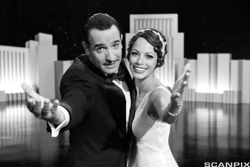 En mann i dress og en kvinne i kjole rekker begge ut ei hånd og smiler til kamera. Svart-hvitt foto.