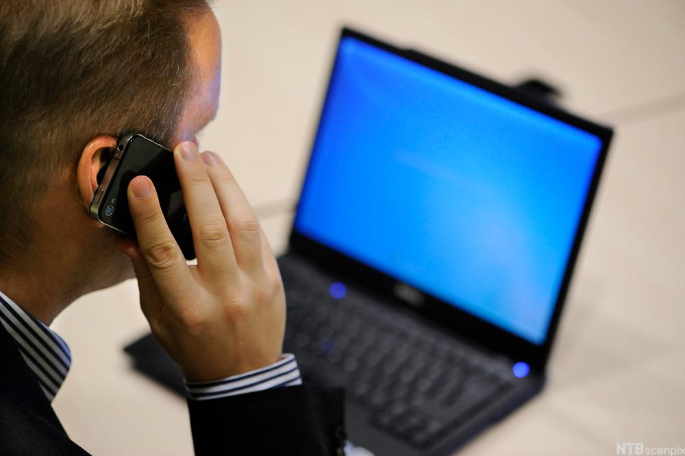 Mann med telefon i hånda og datamaskin foran seg. Foto.