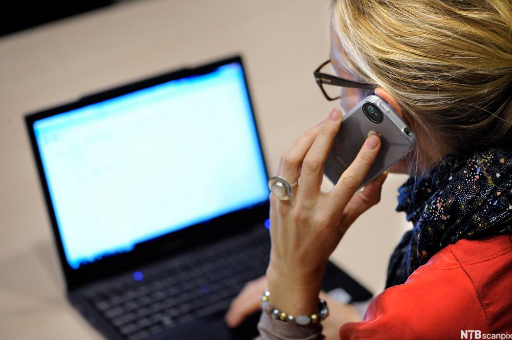 Kvinne sitter ved en bærbar datamaskin og snakker samtidig i telefonen. Foto.