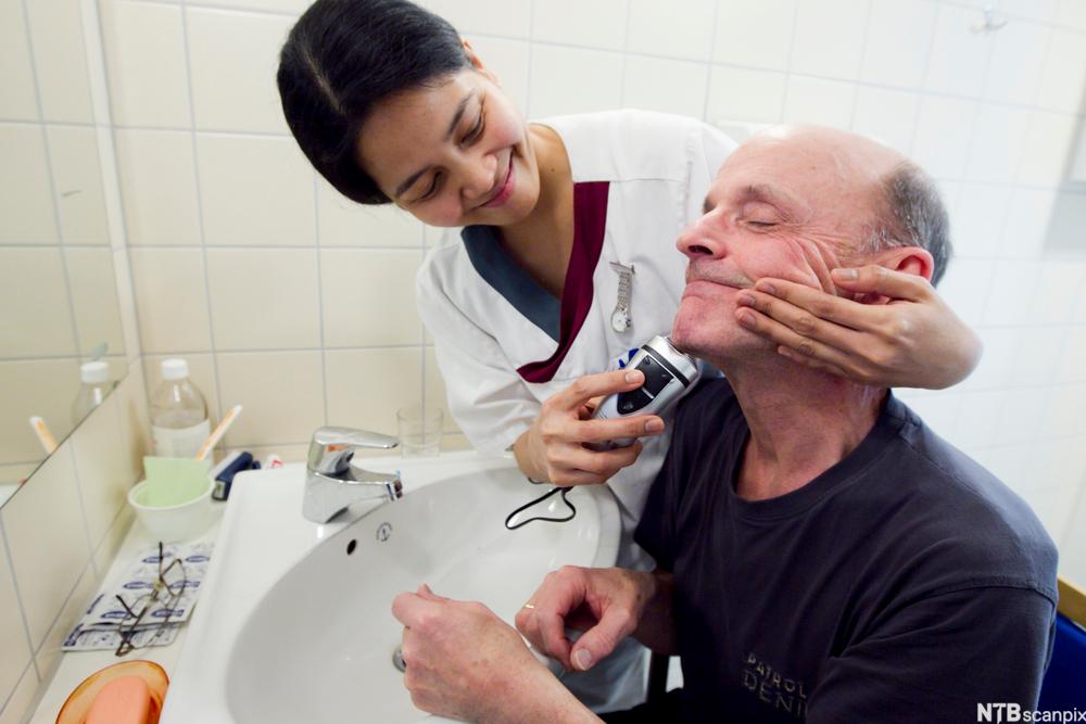 Kvinneleg helsefagarbeidar barberer ein eldre mann med barbermaskin. Foto.