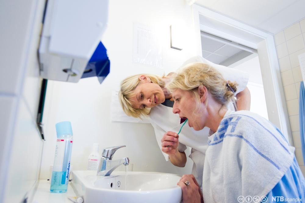 En kvinne får hjelp av en helsefagarbeider med å pusse tennene. Foto.