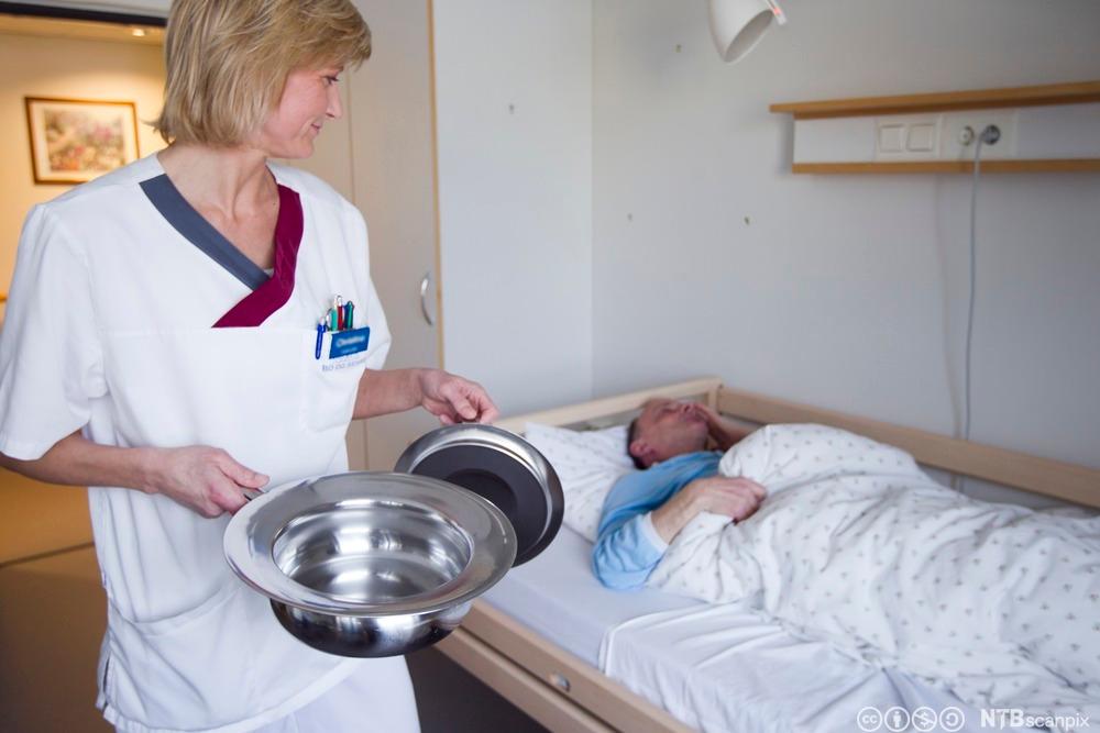Kvinneleg helsefagarbeidar med bekken ved ei seng der det ligg ein eldre mann. Foto.