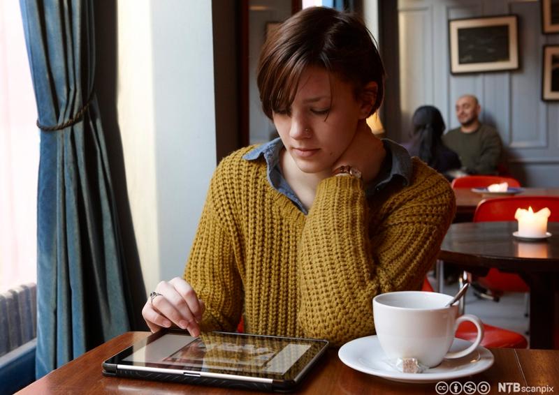 Ung kvinne sitter på kafé og les nyheter på nettbrett. Foto. 