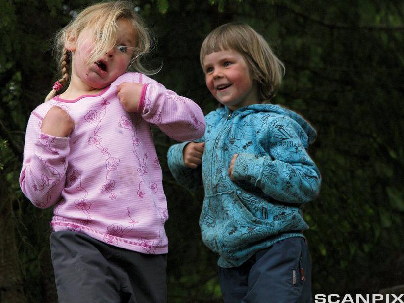 To jenter som går etter hverandre ute i skogen. De har hendene bøyd foran brystet. Den første jenta lager grimaser, den andre jenta smiler. Foto.