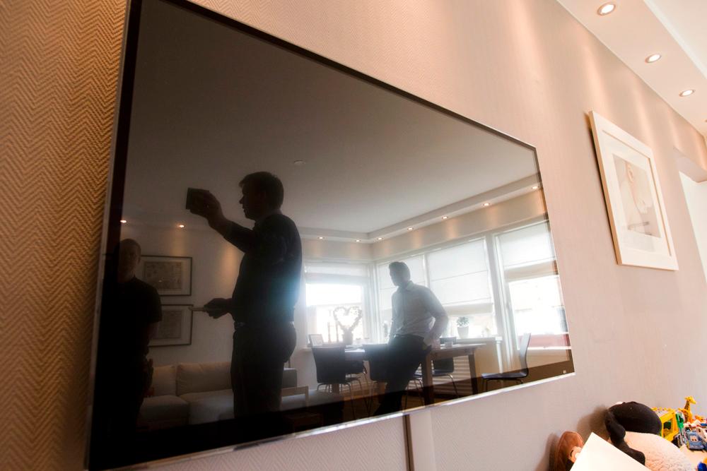 TV-skjerm på en vegg. I speilbildet i skjermen er det tre menn som står og diskuterer noe. Foto.