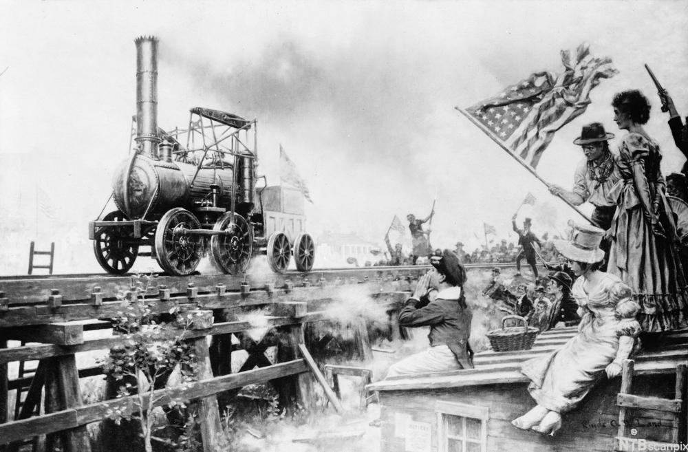Et lokomotiv i USA i 1929. Folk sitter på hustaka og ser på og roper til lokomotivet som går på en skinnegang. En mann vifter med et stort, amerikansk flagg. Bildet har et nasjonalromantisk preg. Foto.