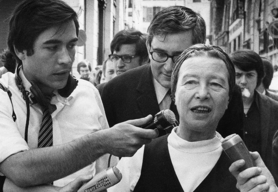 Sartre og de Beauvoir går gjennom ei folkemengde og snakkar med journalistar med mikrofonar. Svart/kvitt-foto.