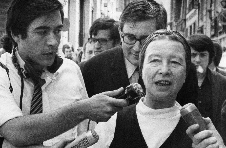 Sartre og de Beauvoir går gjennom en folkemengde og snakker med journalister med mikrofoner. Svart/hvitt-foto.