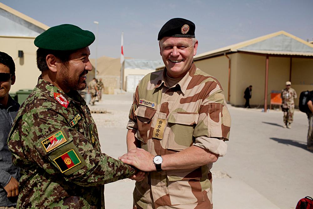 En afghansk og en norsk offiser i kamuflasjeuniformer tar hverandre smilende i hånden i en militærleir i Afghanistan. Foto.
