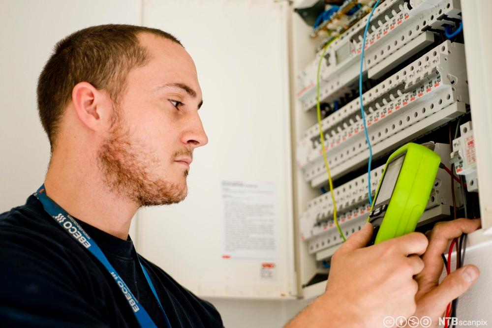 En elektriker sjekker det elektriske systemet i en tavle. Han holder et måleinstrument som blir avlest. Foto.