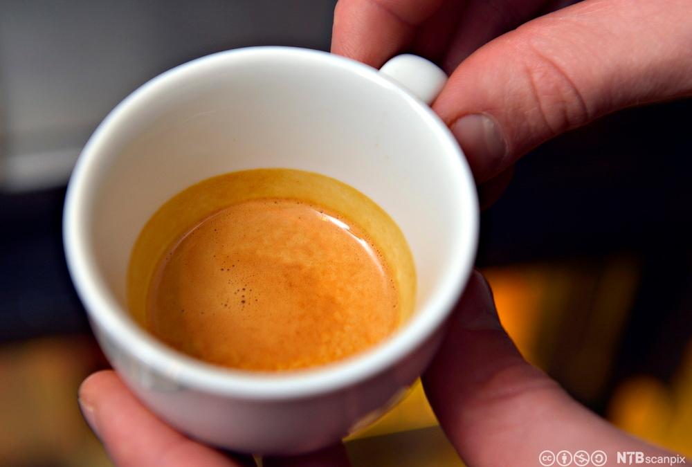 En hånd holder en liten kopp med espresso. Foto.